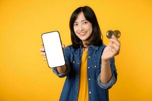 Happy young asian woman wearing yellow t-shirt denim shirt holding digital crypto currency coin and smartphone isolated on yellow background. Digital currency financial concept. photo