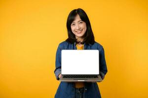 portrait young happy woman wearing yellow t-shirt and denim shirt holding laptop and point finger to screen isolated on yellow studio background. business technology application communication concept. photo