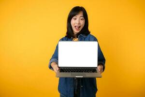 portrait young happy woman wearing yellow t-shirt and denim shirt holding laptop and point finger to screen isolated on yellow studio background. business technology application communication concept. photo