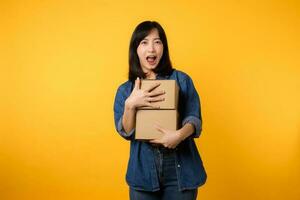 Portrait asian young woman wearing yellow t-shirt and denim shirt holding parcel box isolated on yellow studio background, Delivery courier and shipping service concept. photo