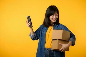 Portrait asian young woman wearing yellow t-shirt and denim shirt holding mobile phone and parcel box isolated on yellow studio background, Delivery courier and shipping service concept. photo