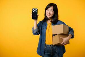 Portrait asian young woman wearing yellow t-shirt and denim shirt holding mobile phone and parcel box isolated on yellow studio background, Delivery courier and shipping service concept. photo