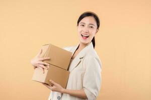 portrait of happy successful confident young asian business woman wearing white jacket holding parcel box isolated on beige studio background, Delivery courier and shipping service concept. photo