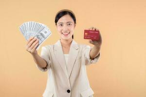 portrait of happy successful confident young asian business woman wearing white jacket holding cash money dollars and credit card standing over beige background. millionaire business, shopping concept photo