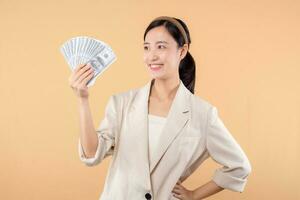 retrato de contento exitoso confidente joven asiático negocio mujer vistiendo blanco chaqueta participación efectivo dinero dolares en pie terminado beige antecedentes. millonario negocio, compras concepto. foto