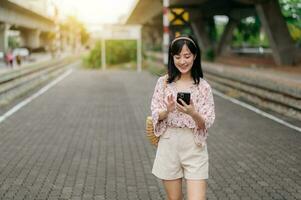 Asian young woman traveler with weaving basket using a mobile phone beside railway train station in Bangkok. Journey trip lifestyle, world travel explorer or Asia summer tourism concept. photo