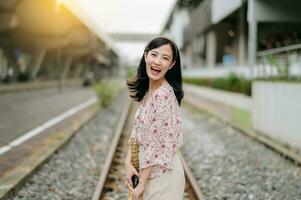 joven asiático mujer viajero con Costura cesta contento sonriente mirando a un cámara junto a tren ferrocarril. viaje viaje estilo de vida, mundo viaje explorador o Asia verano turismo concepto. foto