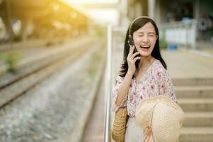 Asian young woman traveler with weaving hat using a mobile phone beside railway train station in Bangkok. Journey trip lifestyle, world travel explorer or Asia summer tourism concept. photo