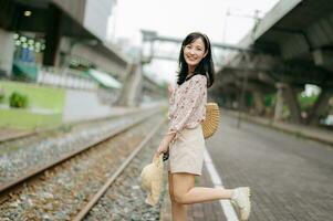 joven asiático mujer viajero con Costura cesta contento sonriente mirando a un cámara junto a tren ferrocarril. viaje viaje estilo de vida, mundo viaje explorador o Asia verano turismo concepto. foto