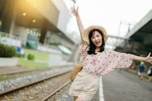 joven asiático mujer viajero con Costura cesta contento sonriente mirando a un cámara junto a tren ferrocarril. viaje viaje estilo de vida, mundo viaje explorador o Asia verano turismo concepto. foto