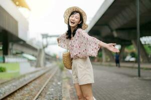 joven asiático mujer viajero con Costura cesta contento sonriente mirando a un cámara junto a tren ferrocarril. viaje viaje estilo de vida, mundo viaje explorador o Asia verano turismo concepto. foto