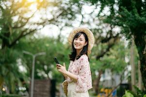 Portrait of asian young woman traveler with weaving hat and basket using mobile phone on green park nature background. Journey trip lifestyle, world travel explorer or Asia summer tourism concept. photo