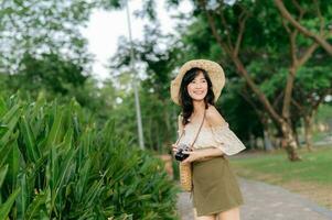 retrato de asiático joven mujer viajero con Costura sombrero y cesta y un cámara en verde público parque naturaleza antecedentes. viaje viaje estilo de vida, mundo viaje explorador o Asia verano turismo concepto. foto