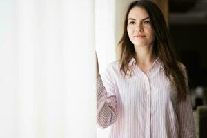 joven hermosa morena mujer a el ventana ligero fondo, toques el cortina con su dedos. foto