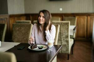 Young beautiful brunette woman sit in coffee shop cafe restaurant indoors and eat chocolate brownie dessert cake. photo