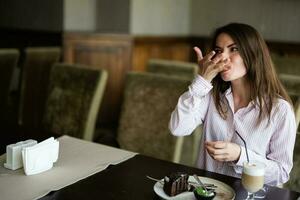 joven hermosa morena mujer sentar en café tienda café restaurante adentro y paliza su dedos mientras comer chocolate duende postre pastel. foto