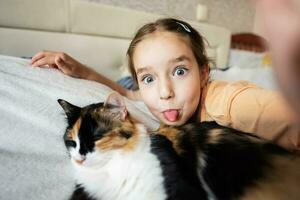 pequeño niña tomando selfie con gato en cama a hogar. retrato de adorable niño espectáculo lengua con mascota. foto