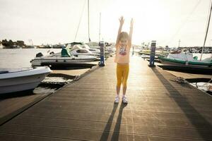 Little girl on a pier with raised hands on sunset boats. photo
