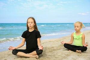 muchachas practicando yoga en playa. niños son sentado en loto posición. foto