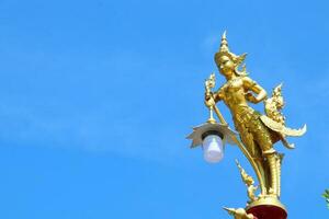 Golden color painted Kinnaree statue holding lamp at the top of pillar and bright blue sky background. Kinnaree is female bird with human head in ancient Thai tale. photo