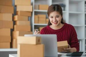 Starting small business entrepreneur of independent Asian woman smiling using computer laptop with cheerful success of online marketing package box items and SME delivery concept photo