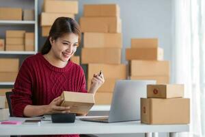 Starting small business entrepreneur of independent Asian woman smiling using computer laptop with cheerful success of online marketing package box items and SME delivery concept photo