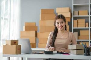 Starting small business entrepreneur of independent Asian woman smiling using computer laptop with cheerful success of online marketing package box items and SME delivery concept photo