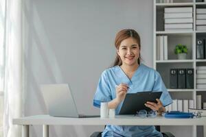 Asian doctor young beautiful woman smiling using working with a laptop computer and her writing something on paperwork or clipboard white paper at hospital desk office, Healthcare medical concept photo