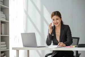 Attractive brunette Asian woman using mobile phone, smile and happy while sitting at working desk in modern office. photo