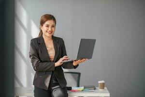 retrato, profesional y confidente asiático mujer de negocios o hembra ejecutivo gerente en formal traje y utilizar ordenador portátil. de pie, propensión en mesa, participación ordenador portátil y utilizando ordenador portátil computadora. foto