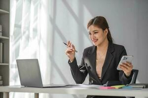 Attractive brunette Asian woman using mobile phone, smile and happy while sitting at working desk in modern office. photo