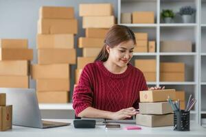 Starting small business entrepreneur of independent Asian woman smiling using computer laptop with cheerful success of online marketing package box items and SME delivery concept photo