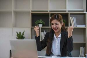 Beautiful Asian businesswoman celebrate while using laptop at office and showing delight. Startup small business and successful concept. photo