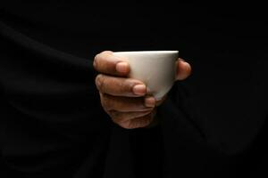 Asian male dark skinned single hand fist finger on black background holding white Japanese Chinese teacup photo