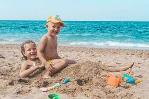 niños hermana y hermano mentira en el playa en el arena. un niña y un pequeño chico jugar en el verano en el mar. verano tiempo. viajar. foto