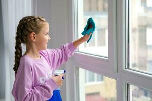 A little girl in a purple sweater sprinkles water from a bottle on the window and wipes it off with a rag. The child helps with cleaning the house. Clean the window. photo