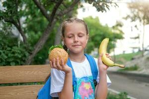 A schoolgirl girl sits on a bench and cannot choose what to eat. Choice of healthy or unhealthy food. Child chooses between burger and banana photo