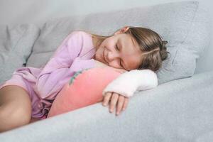 A little girl sleeps on a sofa with a broken arm. The child lies on the couch and put her hand in a cast on the armrest of the couch. photo