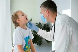 un médico en un médico máscara examina el garganta de un niño. un médico examina un pequeño niña durante un enfermedad. vocación un médico a hogar. foto