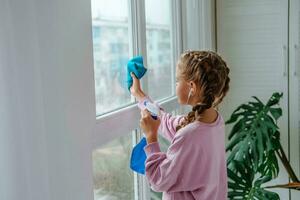 A little girl sprinkles water from a bottle on the window and wipes it off with a rag. The child listens to music on headphones and helps with cleaning the house. Clear the window. photo