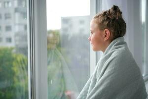 The girl looks out the window at the rain. The child is sad near the window because of the bad weather. photo