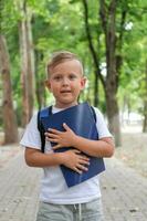 un pequeño chico sostiene un cuaderno en su manos con un mochila en su atrás. el niño es Listo para escuela. foto