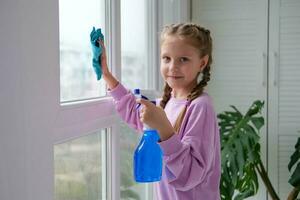 A little girl in a purple sweater sprinkles water from a bottle on the window and wipes it off with a rag. The child helps with cleaning the house and looks at the camera. Clean the window. photo