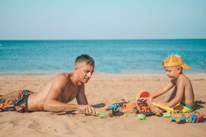 The family is playing on the beach. Son and father are playing with sand on the seashore. Dad with a child on the beach playing with toys. Summer time. Travel. photo