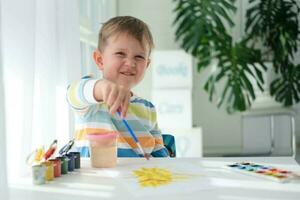 contento niño pintura con cepillo y vistoso pinturas un pequeño chico sorteos un imagen con pinturas en un sábana de papel. el niño es aprendizaje a dibujar. hogar creatividad. foto