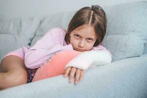 Close-up of a child's broken arm in a cast. The girl put her broken arm on the sofa and looks sadly at the camera. A child with a plaster on his arm. photo