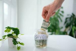 Man's hand puts 1 dollar in a glass jar with the inscription save. Money saving concept. Credit, interest. photo