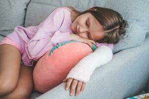 A little girl sleeps on a sofa with a broken arm. The child lies on the couch and put her hand in a cast on the armrest of the couch. photo