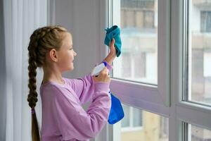 A little girl in a purple sweater sprinkles water from a bottle on the window and wipes it off with a rag. The child helps with cleaning the house. Clean the window. photo