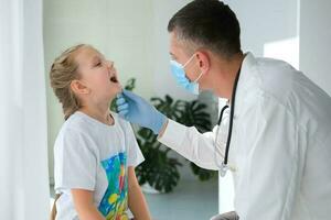 un médico en un médico máscara examina el garganta de un niño. un médico examina un pequeño niña durante un enfermedad. vocación un médico a hogar. foto